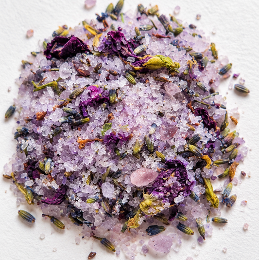 A small pile of foot soak bath salts featuring coarse white crystals mixed with dried lavender buds, blue pea flower buds,   on a textured white background.