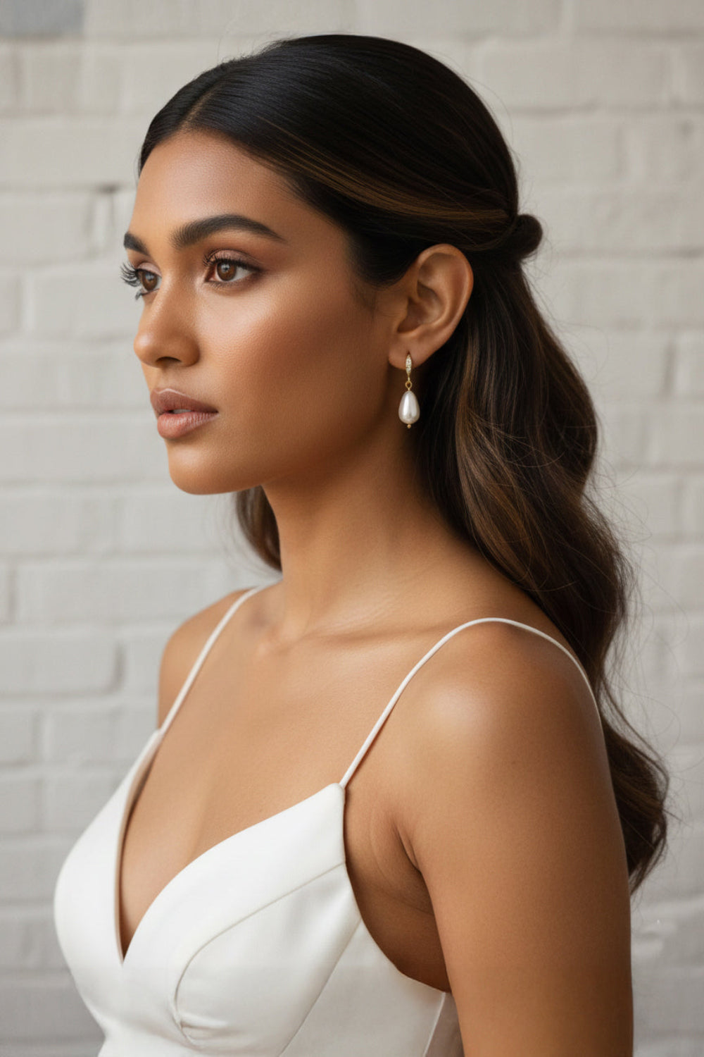 Woman with styled hair and white top against a light brick wall wearing simple elegant pearl teardrop earrings and modern white wedding dress. 