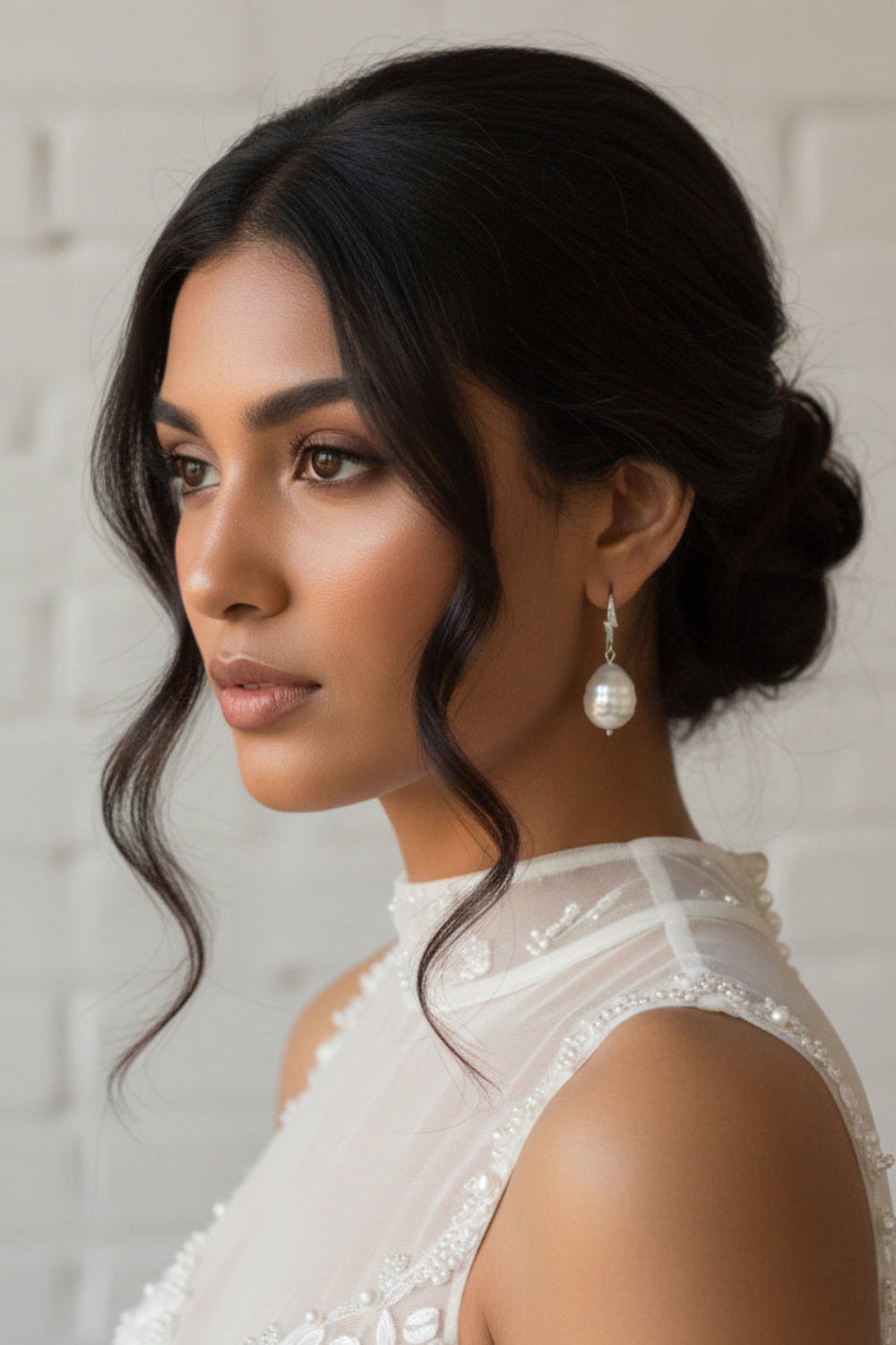 Woman with styled hair and pearl earrings wearing a white lace top against a neutral background